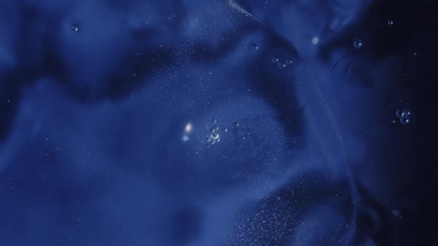 Shimmering particles and air bubbles drift slowly through a viscous deep blue fluid creating soft undulating currents in this macro closeup