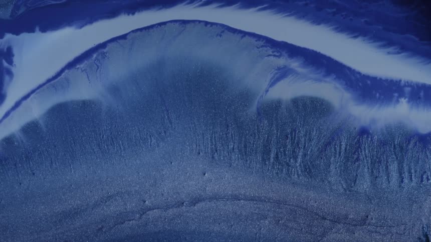 Blue liquid paint flowing across a flat surface with a paintbrush sweeping over it and disrupting the smooth flow creating ripples streaks and shifting currents in the wet layer