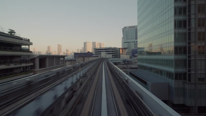 An elevated train track runs straight through a dense urban corridor of modern buildings with the converging rails and surrounding concrete structures creating a strong sense of forward motion and metropolitan scale as the city stretches ahead