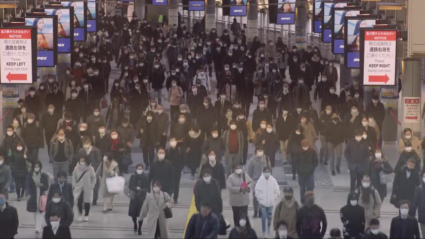 A dense crowd of masked commuters moves in unison through a large brightly lit transit concourse lined with digital screens and directional signs conveying the intensity scale and routine urgency of urban life during a busy rush hour
