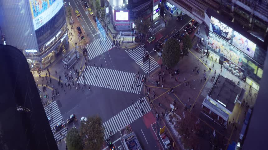 A high angle view captures many people crossing a large city intersection with white painted crosswalks between tall buildings with glowing signs at night
