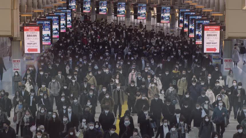 A massive dense sea of commuters almost entirely wearing face masks floods through a wide station concourse in an overwhelming but orderly wave of movement
