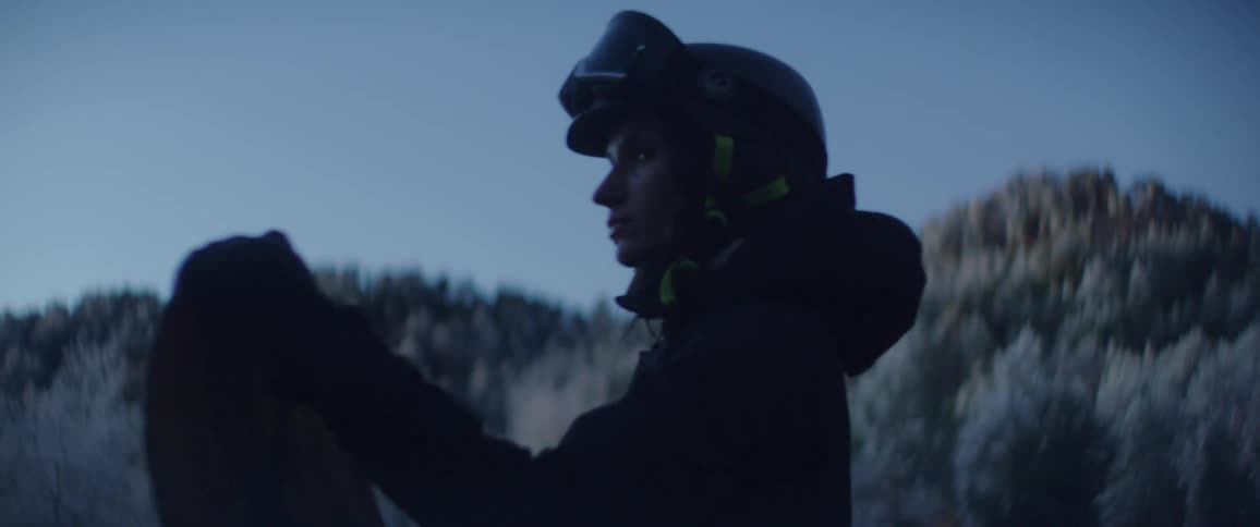 Woman wearing a helmet stands holding a snowboard with snow covered trees on mountains in the distance