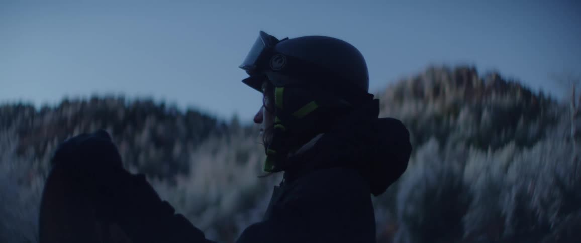 A snowboarder with a helmet and goggles stands on a snowy mountain slope during the blue hour