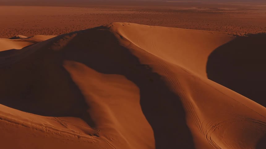An aerial view captures the textured slopes of a massive orange sand dune marked by faint tire tracks and cast in deep shadow against the vast desert horizon