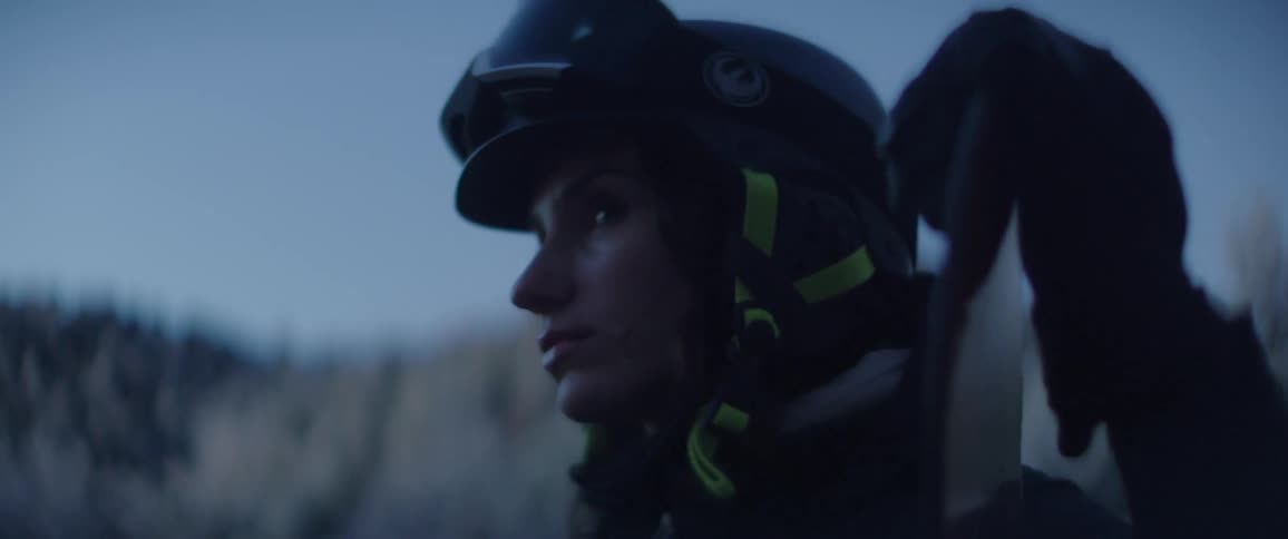 In the cool blue light of dusk a female snowboarder wearing a helmet and goggles rests her board against her shoulder gazing thoughtfully across the winter landscape