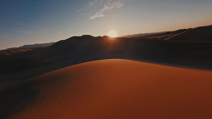 A drone flies over vast sand dunes toward a bright sun setting behind dark mountain ridges