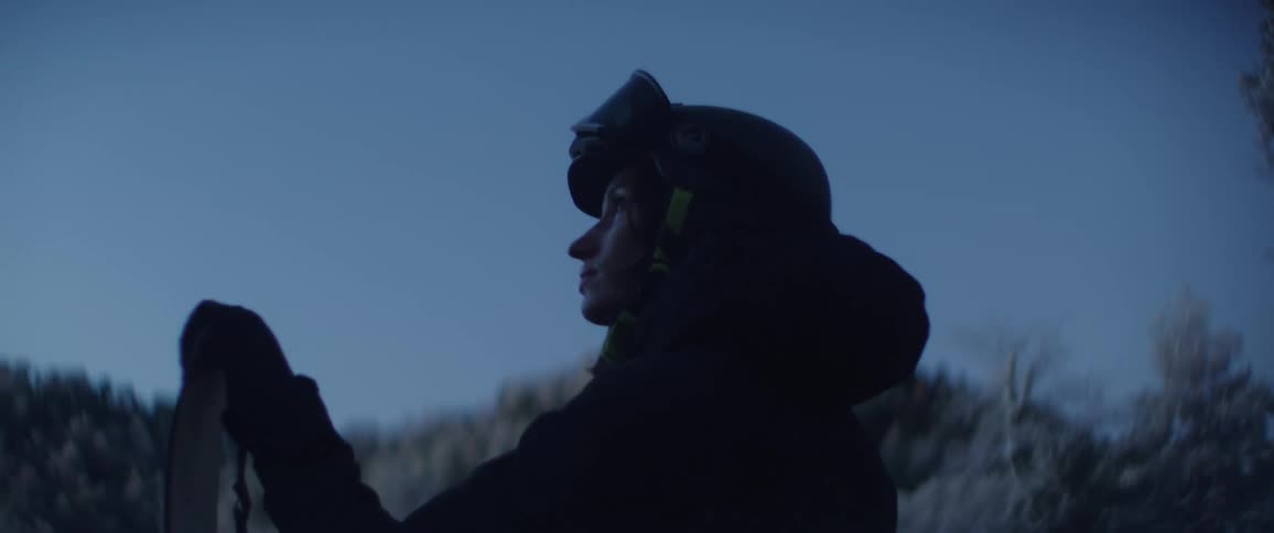 Young adult woman wearing a helmet and goggles holding a snowboard next to a forest outdoors