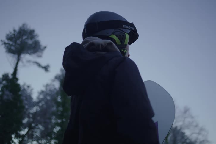 Young adult woman wearing goggles and a helmet standing up next to trees holding a snowboard under her arm outdoors during dusk or dawn