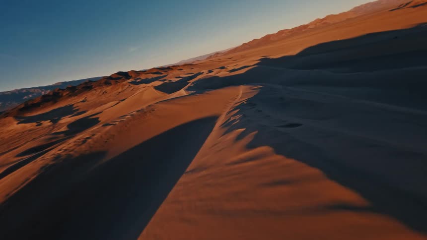 Racing at high speed over the crests of vast sand dunes the view sweeps across the desert landscape where golden sunset light contrasts with deep shadows under a clear blue sky