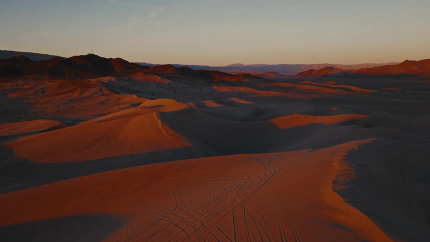 Goldenhour light washes over an expansive desert of rolling sand dunes and distant mountains with long shadows and faint track lines etched into the warm orange surface