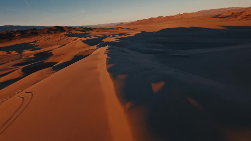 A drone captures a sweeping aerial view of vast sand dunes at sunset gliding low over a sharp ridge while casting long shadows across the desert floor toward distant mountains under a clear blue sky