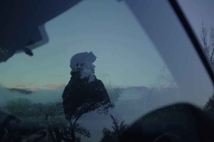 Young female adult seen through a windshield wearing goggles and a helmet looking at a natural landscape with trees and mountains on the background outdoors during dusk or dawn