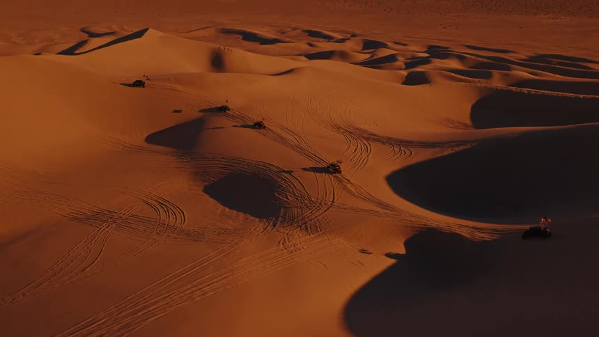 Vehicles drive across sand dunes leaving visible tracks under lowangle light