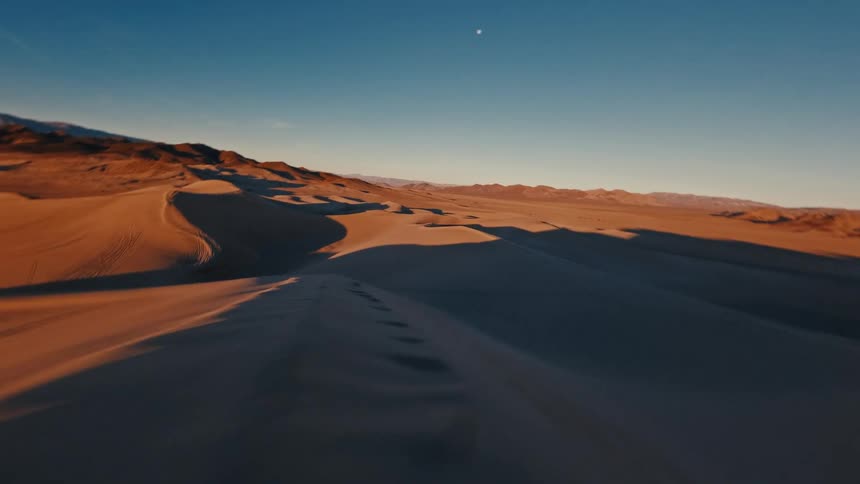 A sweeping aerial perspective glides rapidly over undulating sand dunes marked by tire tracks contrasting the warm sunlit crests with deep shadows beneath a pale moon
