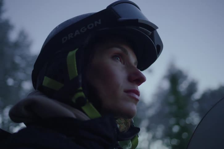 A closeup of a snowboarder wearing a helmet and goggles captures their focused expression as they look upward with cool blue light blurred trees and the edge of the snowboard