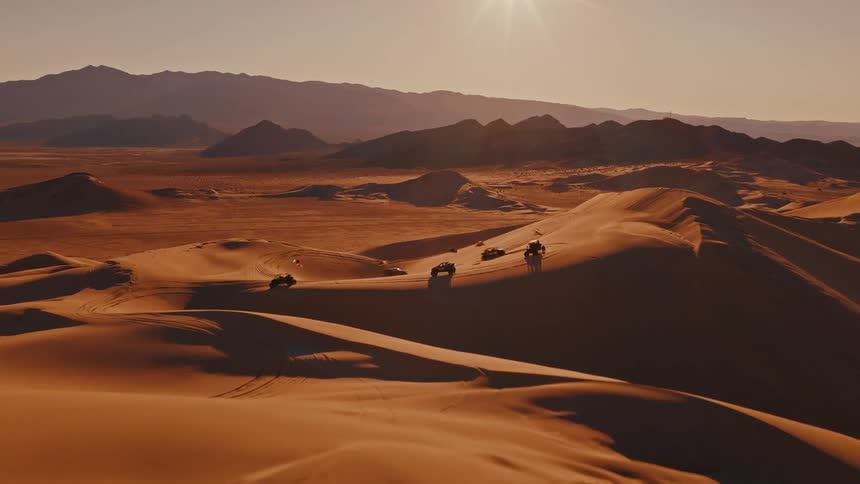 A convoy of offroad vehicles traverses the golden ridges of a vast desert casting long shadows as they speed beneath the warm glow of a setting sun