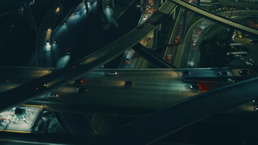 This video features a highangle cinematic drone shot of a complex multilevel highway interchange at night the dark moody atmosphere is punctuated by the glowing headlights and taillights of cars moving through the various overpasses and ramps