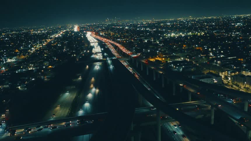 An aerial drone shot pans across a city skyline at night showing heavy traffic flowing along illuminated freeways and sprawling city lights stretching toward the horizon under a dark sky