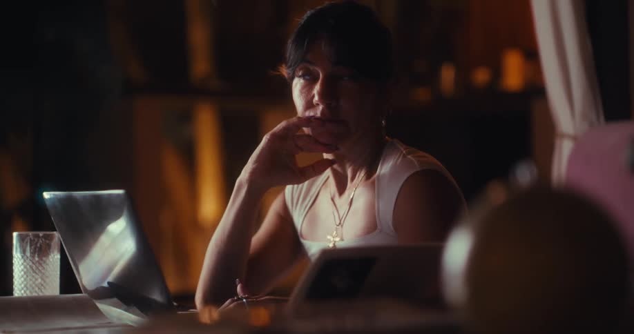 Woman working on a laptop at home at night consults a book