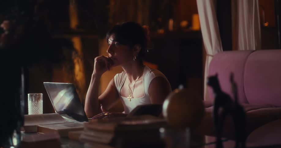 A woman sits at a desk in a dimly lit room resting her chin on her hand while staring thoughtfully at her laptop screen surrounded by books a globe and a pink sofa