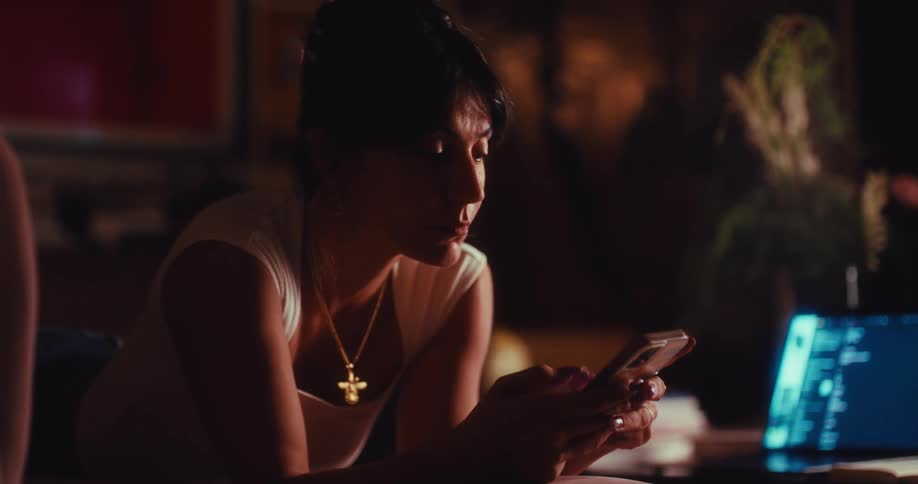 A woman is typing on her cellphone as she sits next to a laptop