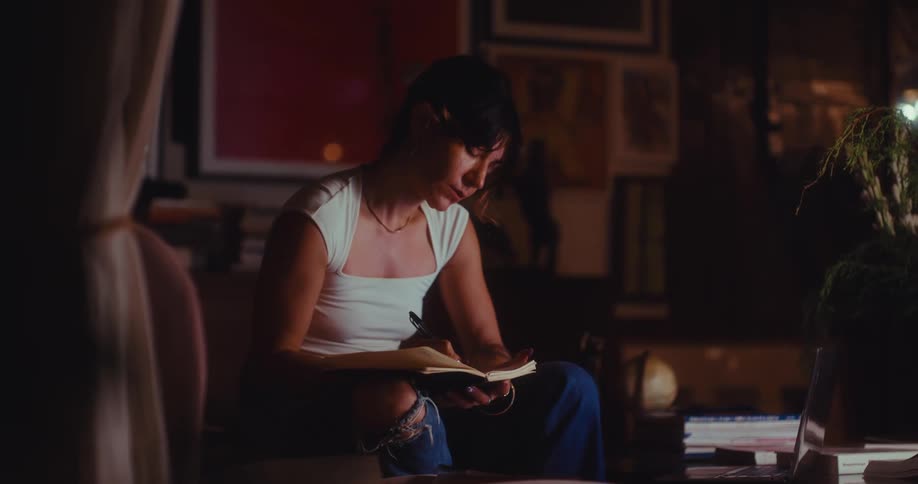 Young adult woman sitting on an armchair and writing on a notebook over her lap with a contemplative facial expression in a living room indoors during night time