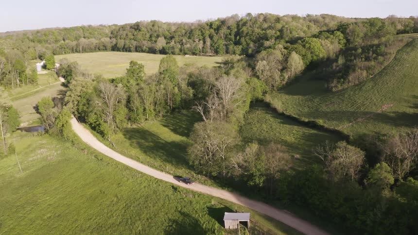 A black car drives along a winding dirt road through a vast rural landscape featuring rolling green hills dense patches of trees and open pastures captured from an elevated aerial perspective during the day