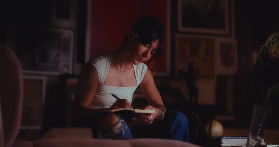 A woman sits in a dark art filled room writing in a notebook and she pauses to tap a pen against her chin while looking away in thought before returning to her journal surrounded by framed sketches and studio equipment in the background
