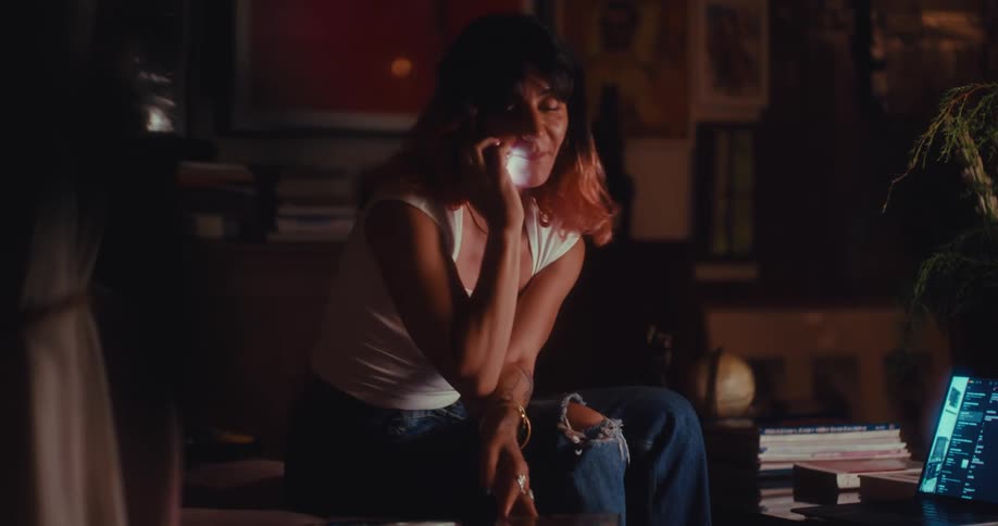 A young woman sitting on a chair in her living room smiling as she answers a phone call with a laptop open on the coffee table in front of her