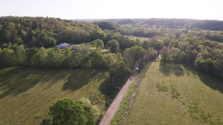 An aerial camera tracks a pickup truck driving along a narrow dirt road through rolling green farmland gradually descending as the vehicle approaches a distant house nestled in the trees