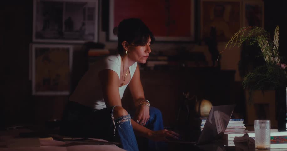 The woman sits down and opens her laptop then picks up a notebook