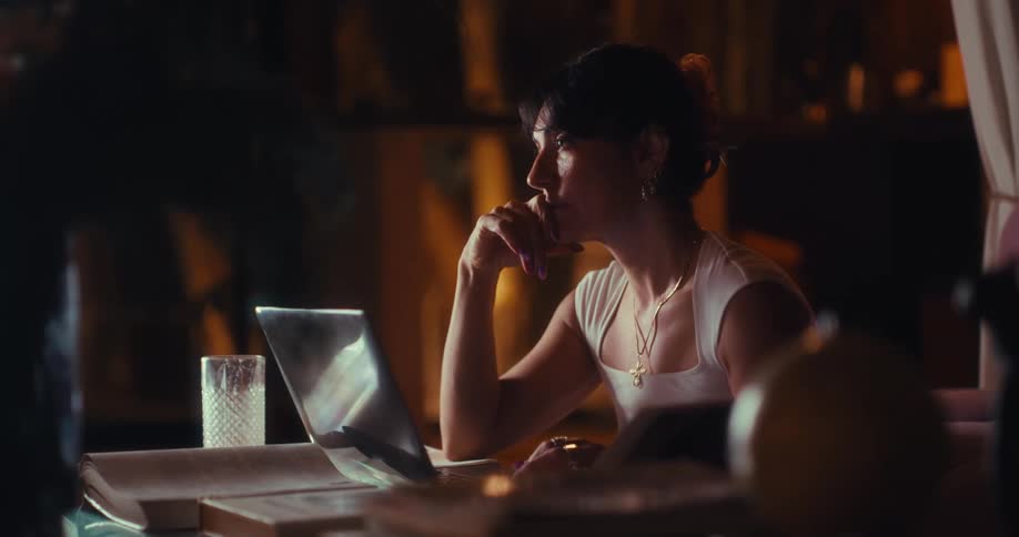 A young woman typing on a laptop in a dimly lit living room seated at a desk with a glass of water placed beside the laptop