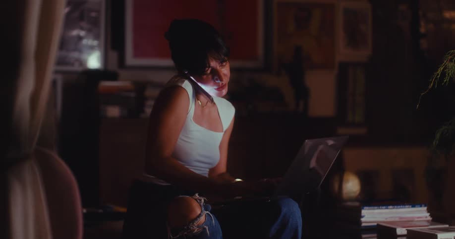 Young woman sitting in a living room chair as she makes a phone call and types on a laptop computer