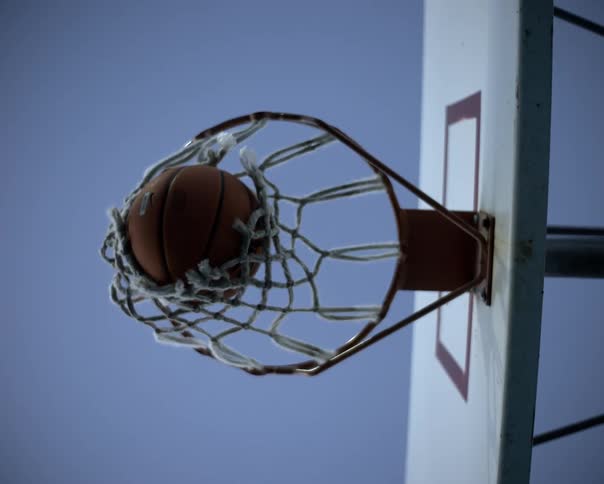 A basketball dropping through the hoop at midday slipping out of the net and bouncing down toward the court floor