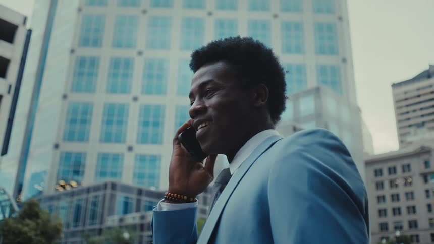 A man in a blue suit smiles while talking on his phone standing in front of a tall glass building
