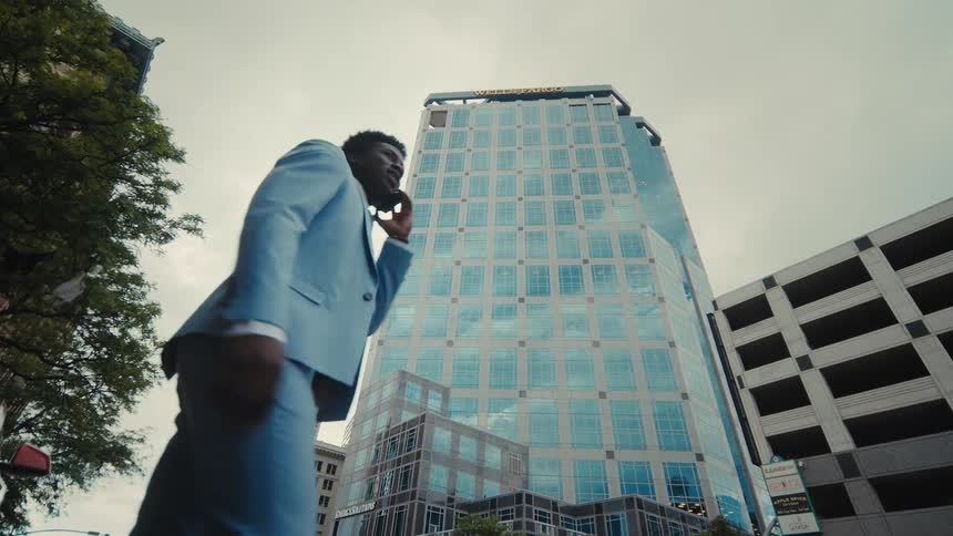 A man in a light blue suit walks on a city sidewalk and looks up at a tall glass skyscraper while talking on his mobile phone