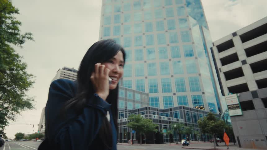 An asian woman in a blue blazer talks on her smartphone while walking along a city street lined with modern glass skyscrapers parked cars and urban greenery under an overcast sky