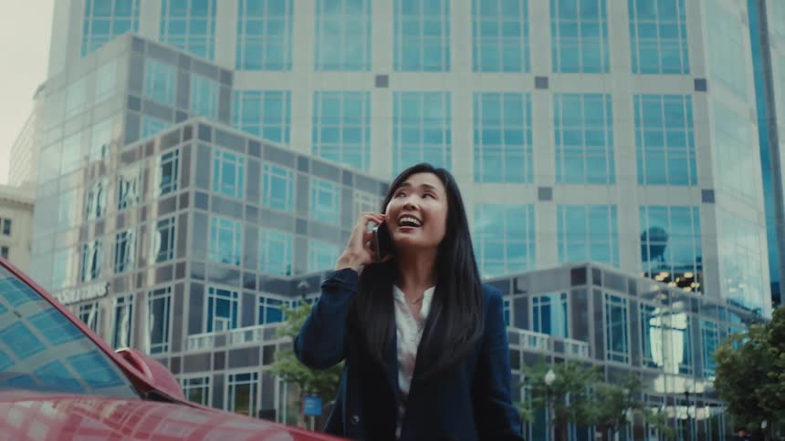 A professional woman talks on her smartphone and smiles while walking past a red car in a city plaza with a large glass office building in the background
