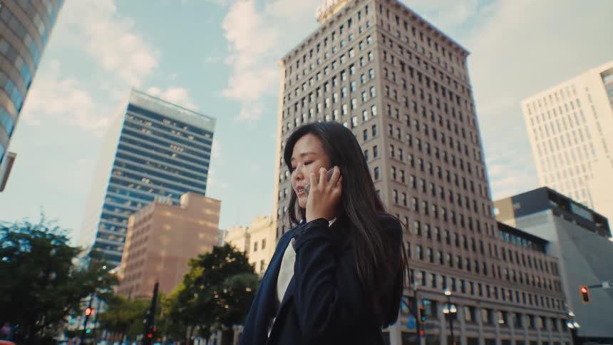 A professional woman engages in a phone conversation at a vibrant city intersection framed by tall skyscrapers and the soft glow of afternoon light