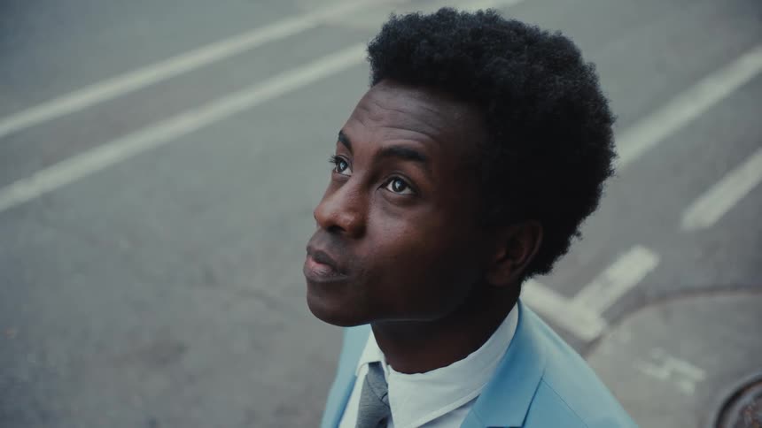 A man wearing a light blue suit and a tie stands on a city street and looks upward with a serious expression