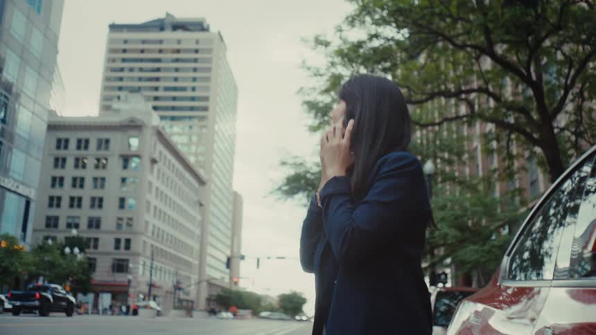 A woman stands on a busy city street talking excitedly on her phone while leaning against a red carshe then smiles broadly and begins to walk away from the vehicle looking happy as she continues her conversation