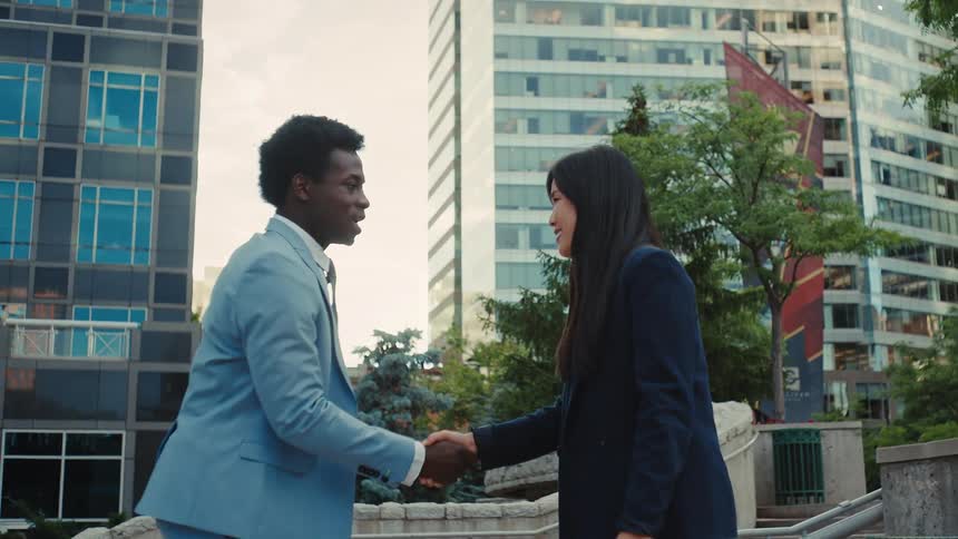 Two professionals shake hands in an outdoor urban setting framed by modern office buildings and greenery