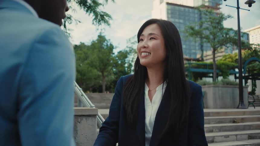 Adult asian woman walking towards an adult black man and smiling while shaking his hand on a staircase at a park in a metropolitan area outdoors during day time