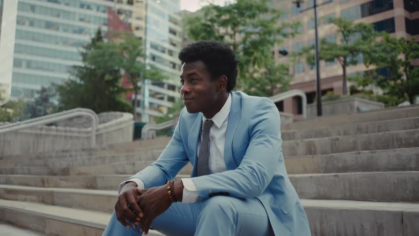 A man wearing a light blue suit and a tie sits on concrete steps in a city plaza with tall buildings and green trees in the background