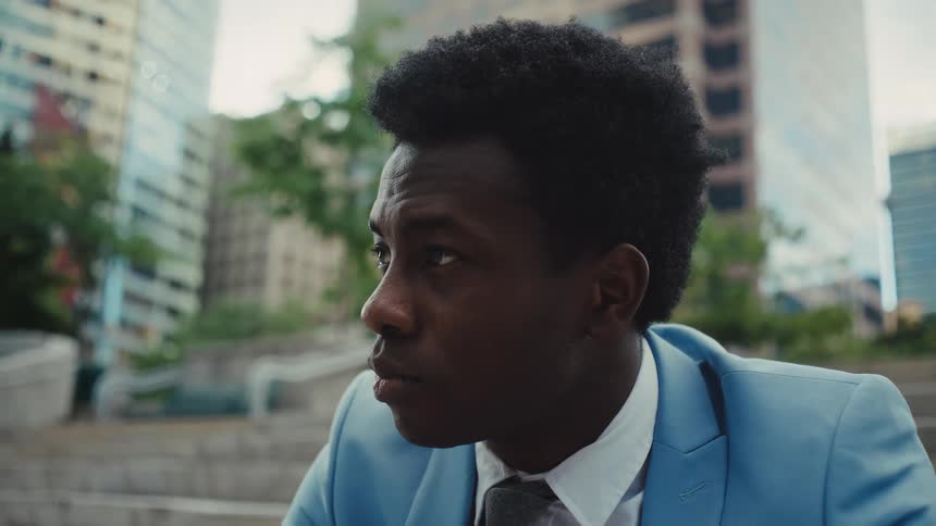 Young adult black man wearing a blue suit and a tie sitting on the staircase of a park in a metropolitan area outdoors during day time
