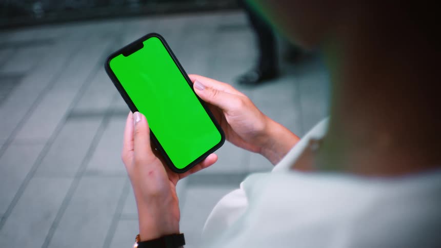 A person holds a smartphone with a bright green screen while standing on a tiled floor