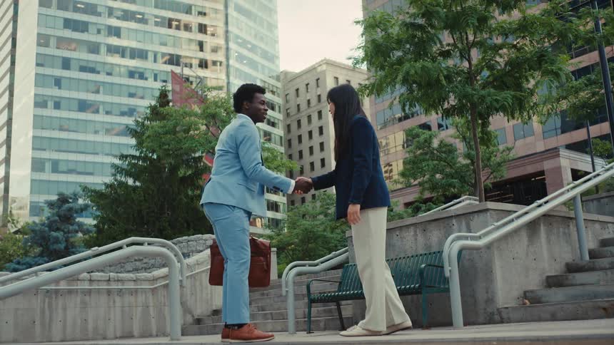 A man in a light blue suit and a woman in a navy blazer meet on a set of concrete stairs to shake hands and exchange a quick greetingafter their brief interaction they both turn and continue walking up the stairs together toward the surrounding city b