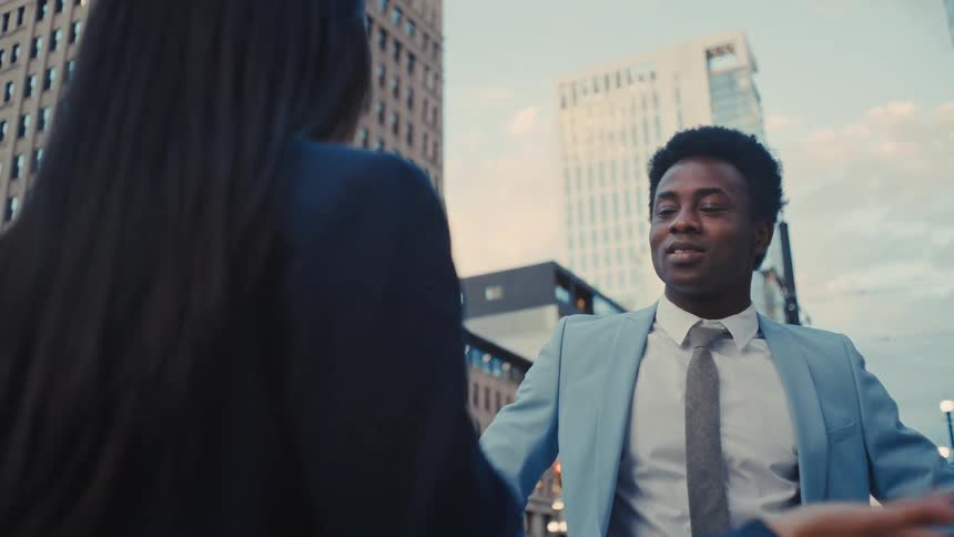 Young adult black man wearing a blue suit and a necktie standing up on the streets of a metropolitan area talking to a woman dressed in black outdoors during day time