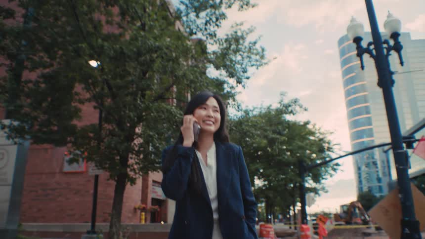 Woman smiles and turns around as she walks on a city sidewalk talking on her phone